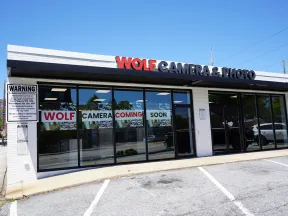 Storefront of Wolf Camera & Photo with signs announcing new store coming soon under clear blue sky.
