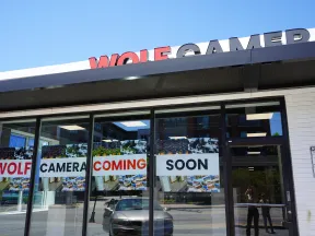 Storefront with signs in windows announcing Wolf Camera coming soon under a clear blue sky.