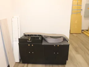 Black printer or copier machine in a room with wooden floor and white walls, surrounded by stacked boxes and panels