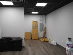 Empty room with light wood flooring, gray walls, black ceiling tiles, cardboard boxes, and paint supplies on the floor.