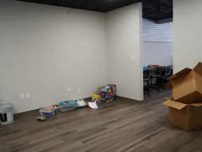 Empty office corner with unpacked boxes, paint buckets, and plastic containers on wooden floor near white walls.