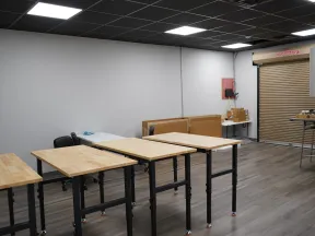 Empty workshop room with wooden tables, black chairs, gray walls, and a roll-up door on wood flooring.