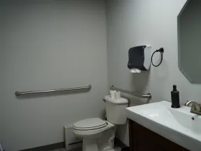 Accessible public restroom with toilet, sink, handrails, paper towel dispenser, and mirror in a clean, simple layout.