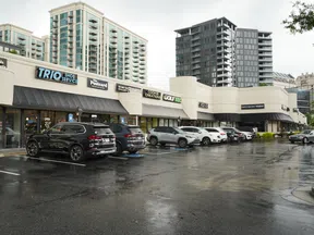 Shopping center with parked cars on wet pavement under cloudy sky in urban area with high-rise buildings.