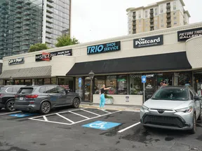 Street view of small retail shops including Trio Shoe Service, The Postcard cafe, and parking with cars and a person in bright pants.