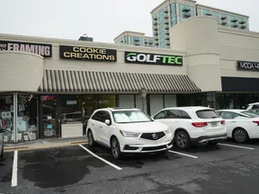 Strip mall storefronts with shops named Cookie Creations, GolfTec, and Moda 404 with cars parked in front on a cloudy day