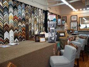 Interior of a frame shop with wall display of frame corners, workstations, and seating area with upholstered chairs.
