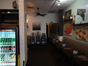 Cozy cafe interior with stacked chairs, wall art, cushioned bench seating, and a refrigerated display of beverages.
