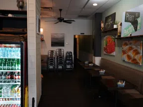 Interior of a small restaurant with stacked chairs, tables, wall art, and a refrigerator of bottled drinks.
