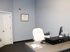 Modern office with a wooden desk, white leather chair, dual monitors, and framed certificate on light gray wall.