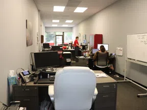 Modern office space with desks, computers, whiteboard, and employees working in an open layout under fluorescent lighting