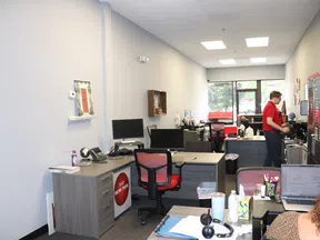 Modern office space with desks, computer monitors, office chairs, and a person working near the window.
