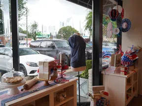 Patriotic-themed shop window with blue dress on mannequin, gift boxes, decorations, and baked goods with city street background