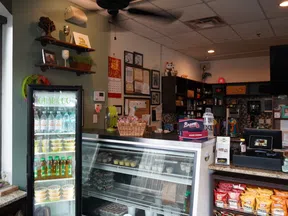 Interior of a small deli with refrigerated display cases, snacks, certificates on wall, and cash register counter.