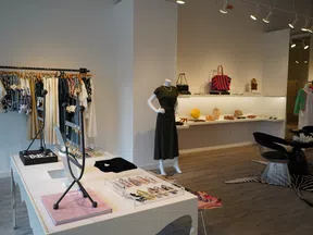 Modern boutique interior featuring clothing racks, accessories display, mannequin with black dress, and seating area.