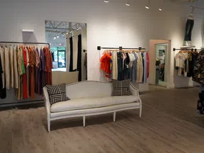 Bright clothing store interior with wooden floor, racks of colorful clothes, and a white cushioned sofa with patterned pillows.