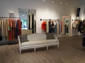 Bright boutique interior with clothing racks, a white vintage sofa, and wooden flooring under soft lighting