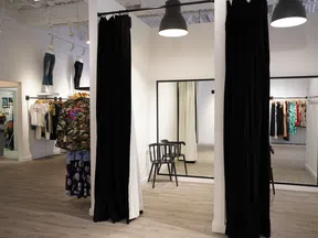 Clothing store dressing room with black curtains, wooden chair, large mirrors, and hanging apparel on racks.