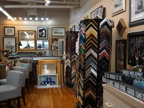 Interior of a frame shop with various frame samples, framed art, mirrors, and decorative signs on wooden floor.