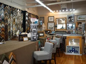 Interior of a frame shop displaying various picture frames, mirrors, and framed artworks on walls and tables.