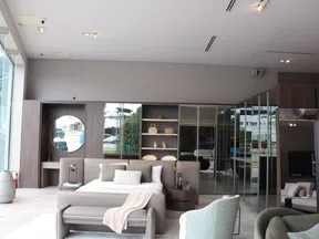Modern bedroom showroom with neutral tones, bed, armchairs, round mirror, and decorative shelving under bright lighting.