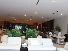 Modern office lounge with white sofas, green plants, and workstations in a bright, spacious room.