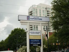 Outdoor multi-business sign for PGA Tour Superstore, Men's Wearhouse, Icebox, Verizon with city buildings and trees.