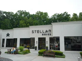 Stellar Bodies retail storefront with benches, potted plants, large glass windows, and green trees in the background.
