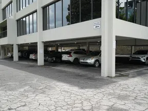 Covered parking lot under an office building with various cars parked and cracked asphalt pavement