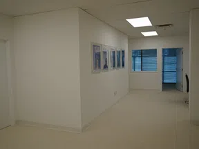 Bright office hallway with white walls, ceiling lights, framed pictures, and a window overlooking urban buildings.