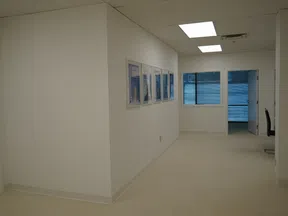 Empty office hallway with white walls, framed photos, fluorescent ceiling lights, and a window at the end.