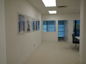 Bright office hallway with framed photos on wall and windows with blue blinds leading to rooms with chairs and tables.