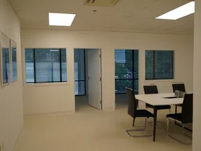 Bright, empty office room with beige carpet, white walls, table with black chairs, and windows showing outdoor greenery.
