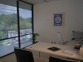 Modern office with large window blinds, white desk, black chair, and a green plant by the window.