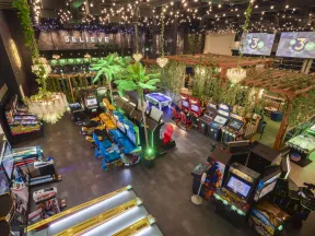 Indoor arcade with vintage and modern game machines, colorful seats, green plants, and string lights above.