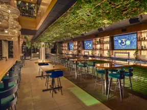 Modern bar interior with green ceiling plants, wooden tables, blue chairs, and illuminated shelves with bottles.