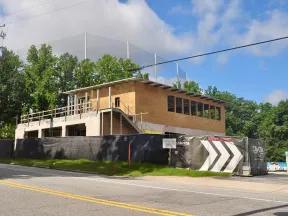 Two-story building under construction with plywood walls and black fence, surrounded by trees and clear sky.