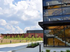 Modern glass building with visible staircases beside a green lawn and partly cloudy blue sky.