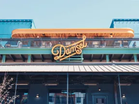 Outdoor view of Damsel restaurant with illuminated sign and rooftop seating under a clear blue sky.