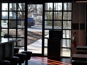 Interior view of a cafe with stools and tables near large glass windows showing an outdoor sidewalk and passing car.