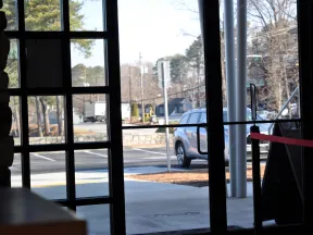View through glass doors showing a parking lot, a parked silver SUV, and trees on a sunny day.