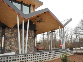 Modern building exterior with angled wooden roof, white pillars, stone wall, and outdoor ceiling fans overlooking a garden.