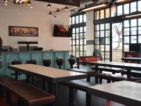 Modern indoor bar with long tables, leather benches, bar stools, large windows, and industrial lighting fixtures.