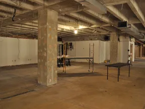 Empty unfinished basement with exposed concrete pillars, ceiling beams, and construction equipment scattered around.
