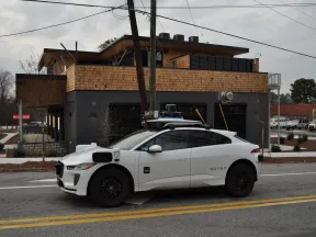 White Waymo self-driving electric car parked on a city street with modern building in the background.