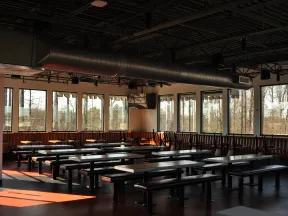 Empty modern cafeteria with large windows, wooden benches, and overhead ventilation ducts during daytime.