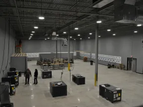 Large industrial warehouse with HVAC units, worktables, and two people standing on concrete floor under high ceiling lights.