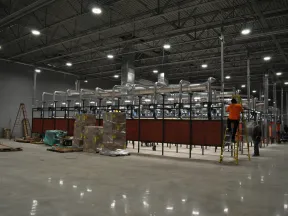 Large industrial warehouse with machinery, ventilation ducts, pallets, and workers performing maintenance tasks.
