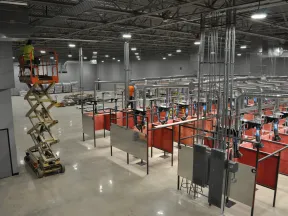 Worker on scissor lift inside large industrial workshop with welding stations and ventilation system.
