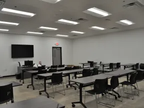 Spacious modern classroom with black chairs and tables, ceiling lights, TV screen, and white walls.
